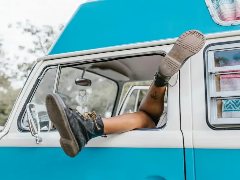 A person's feet, wearing black boots, relaxing out of the window of a blue and white camper van.