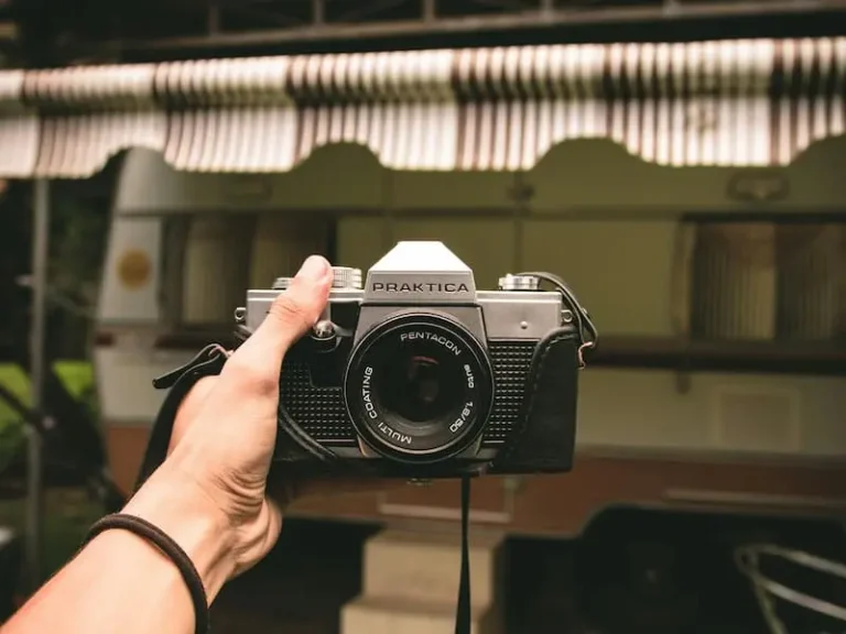 A person's hand holding a vintage film camera, ready to take a photo in front of a caravan.