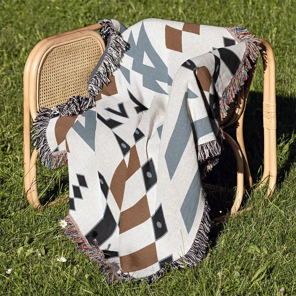 The Wandering Warrior Blanket draped over a light brown rattan chair on a sunny green lawn, showing the fringe detail.