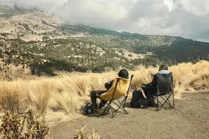 Two campers resting on portable camping chairs, overlooking a vast, scenic mountain range covered in pine forests under a cloudy sky.
