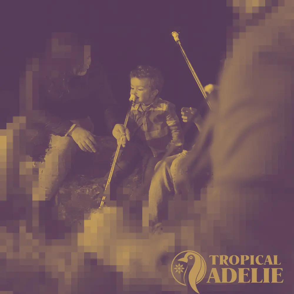 A young child sits by a campfire at night, biting into a piece of damper or a marshmallow on a long stick held by an adult. The image has a warm, purple and gold monochromatic filter with a pixelated aesthetic. In the bottom right corner, a logo for "Tropical Adelie" is visible, featuring a stylized penguin.