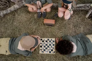 A high-angle shot of four children in scout-like outdoor gear gathered around a wooden radio and a chessboard on a grassy campsite. Two children lie on the grass playing chess, while two others sit nearby against a fallen log.