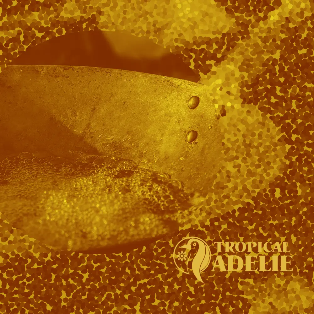 A close-up view of a golden-yellow liquid bubbling and boiling inside a large metallic vat or pot. The scene is framed within a circular vignette against a textured, golden mosaic background. The 'Tropical Adelie' logo, featuring a stylized penguin, is in the lower right.