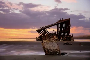 The rusted iron skeleton of the Peter Iredale shipwreck on a wide, sandy beach at sunset, with purple and orange clouds in the sky.