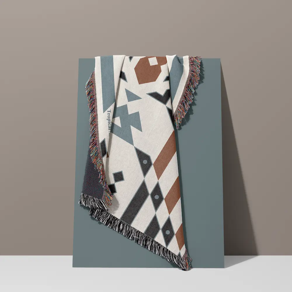 Wandering Warrior woven cotton blanket draped against a slate blue panel in a studio setting, showcasing a bold geometric pattern in natural white, slate blue, warm terracotta and charcoal, with multicolour fringe edges.