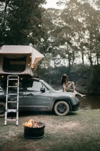 Woman camping alone beside a river, sitting on top of a jeep with a rooftop tent and string lights, ladder leaning against the vehicle, campfire burning in the foreground, surrounded by trees at dusk