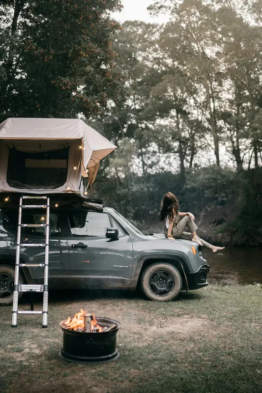 Woman camping alone beside a river, sitting on top of a jeep with a rooftop tent and string lights, ladder leaning against the vehicle, campfire burning in the foreground, surrounded by trees at dusk