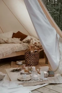 A cozy glamping tent interior in the forest with a wooden bed, layered blankets, candles, and dried flowers in a wicker basket.