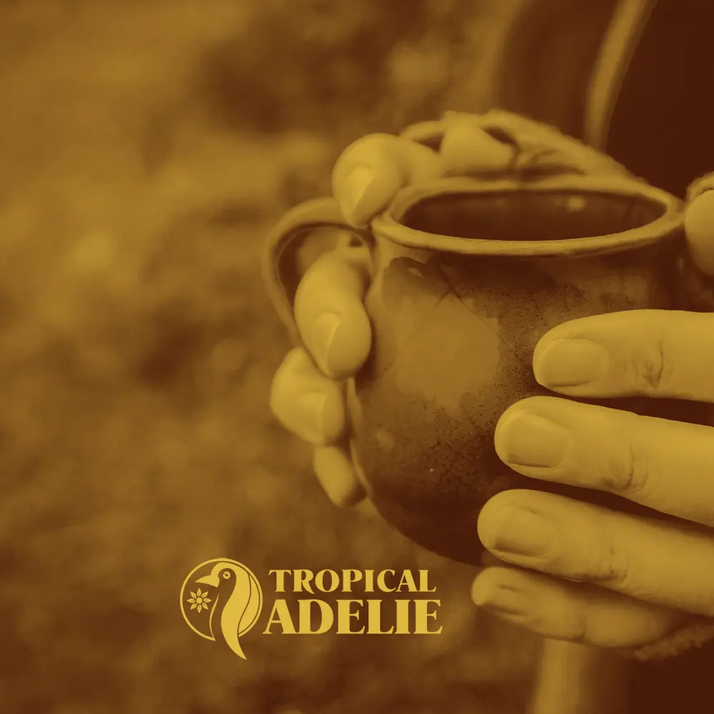 A warm, monochromatic sepia-toned close-up image showing a pair of hands closely holding a ceramic mug against a blurred natural background. The yellow "TROPICAL ADELIE" penguin logo and text are printed at the bottom center.