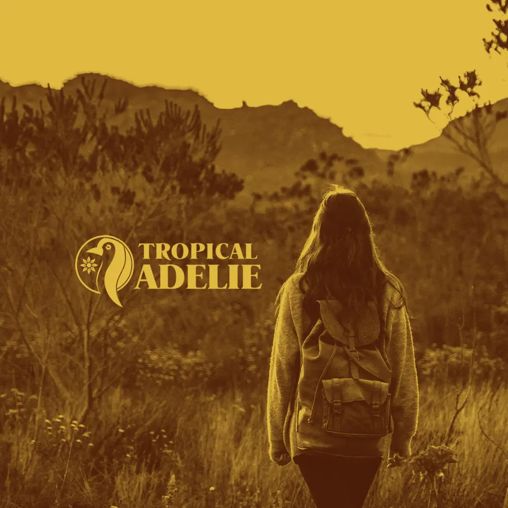 A sepia-toned scenic photograph taken from behind a woman with long hair wearing a knit sweater and a vintage canvas backpack. She is walking through a grassy field towards rolling mountains in the distance. The yellow "TROPICAL ADELIE" penguin logo and text are positioned on the middle-left side.