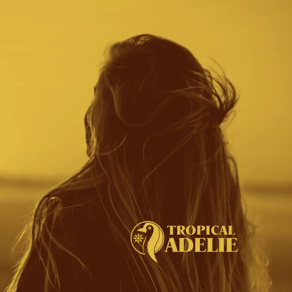 A warm golden-toned close-up shot from behind a person, focusing on their long, wind-blown hair against a soft, brightly lit background. The yellow "TROPICAL ADELIE" penguin logo and text are placed in the bottom right corner.