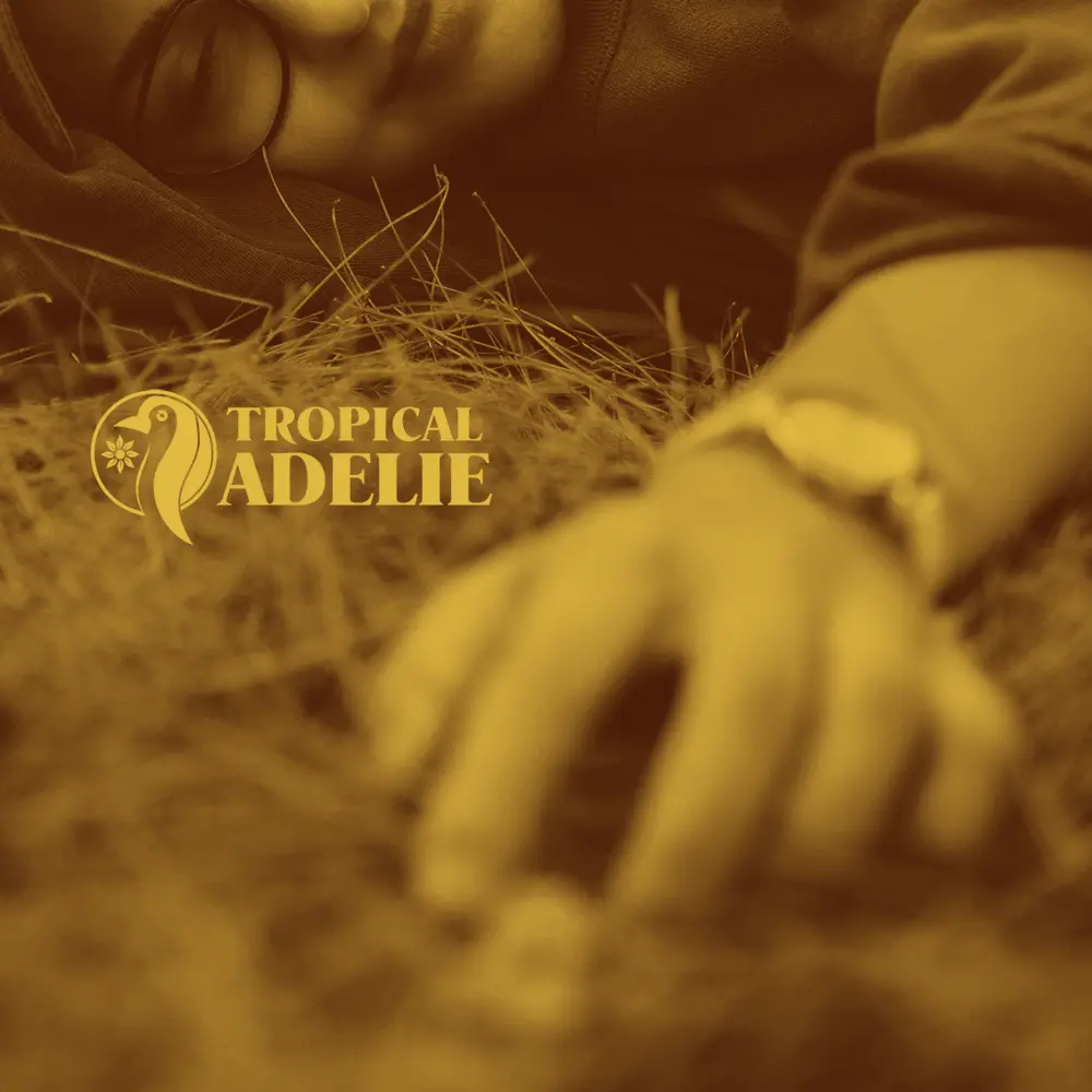 A sepia-toned close-up shot with a shallow depth of field. In the foreground, a blurred hand wearing a wristwatch rests on a bed of dry grass. In the background, the upper portion of a person's face is visible, lying down with closed eyes. The yellow "TROPICAL ADELIE" penguin logo and text are placed on the left side.