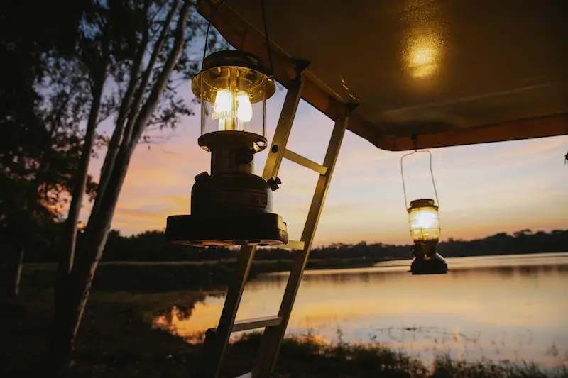 Two glowing oil lanterns hanging from a camp shelter frame at dusk, with a calm lake and golden sunset sky in the background.