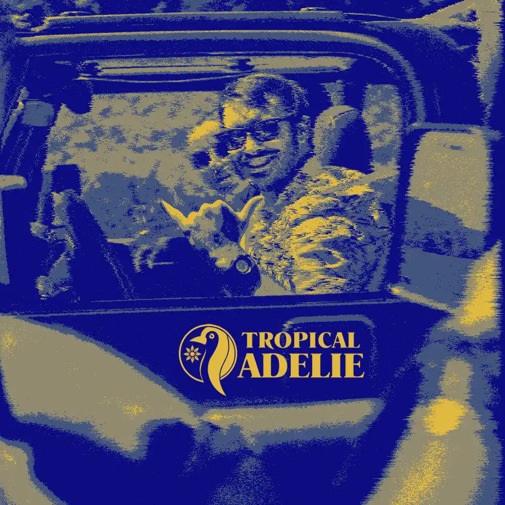 A smiling person sitting in a car with a dog, photographed in a blue and gold duotone style with the Tropical Adelie logo overlay.