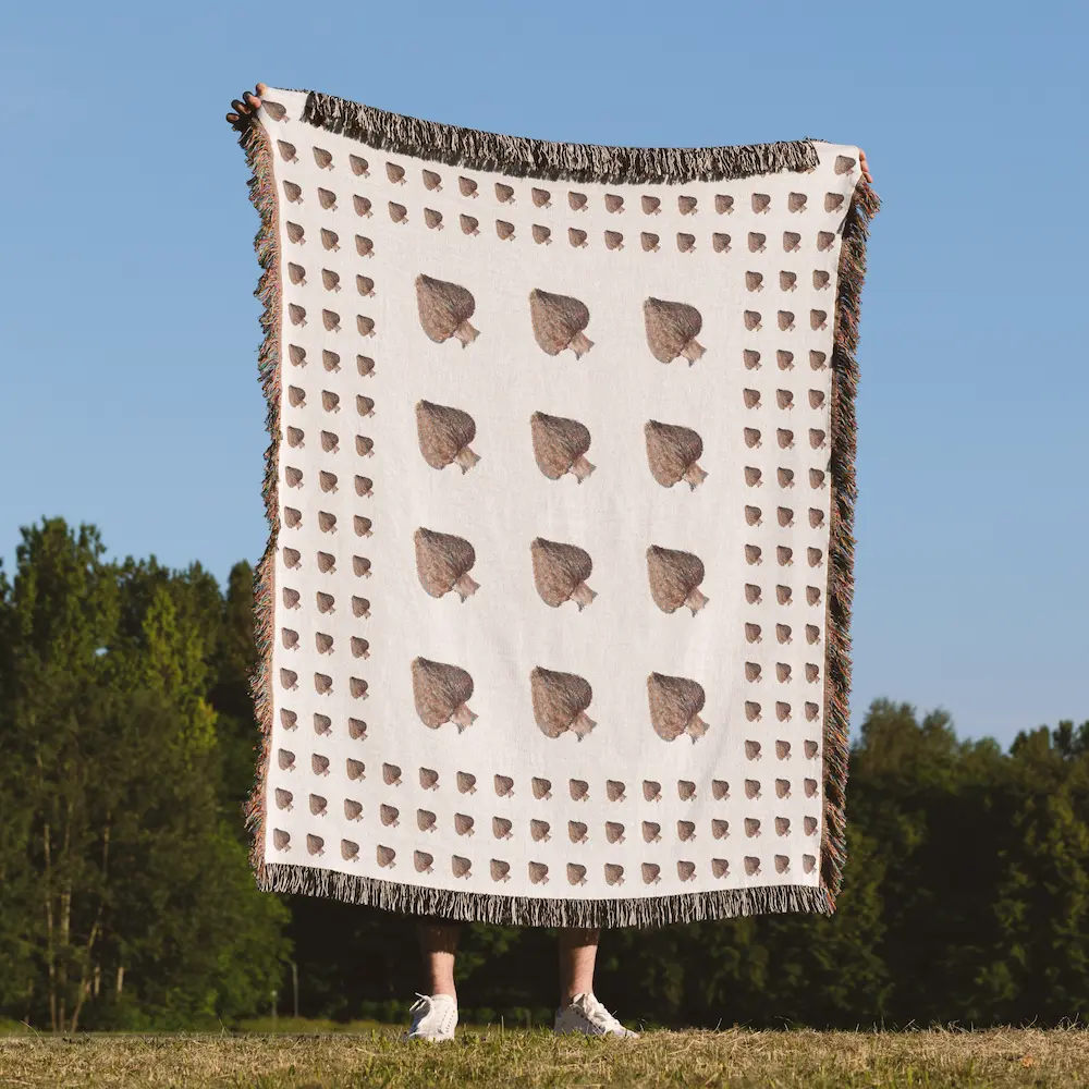 A person holding up the full blanket outdoors against a blue sky and tree line, displaying the mushroom pattern across the fabric.