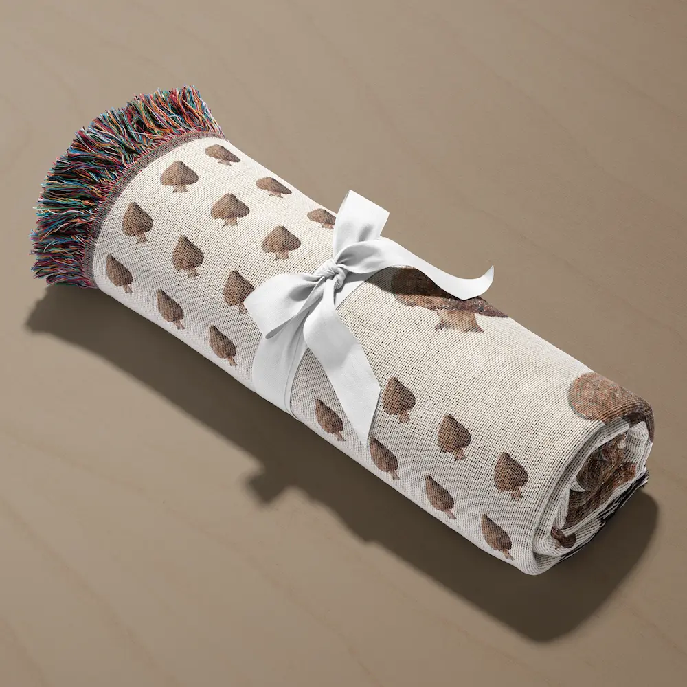 Blanket rolled into a cylinder and tied with a white ribbon, resting on a beige surface, showing the mushroom pattern and dark fringe edge.