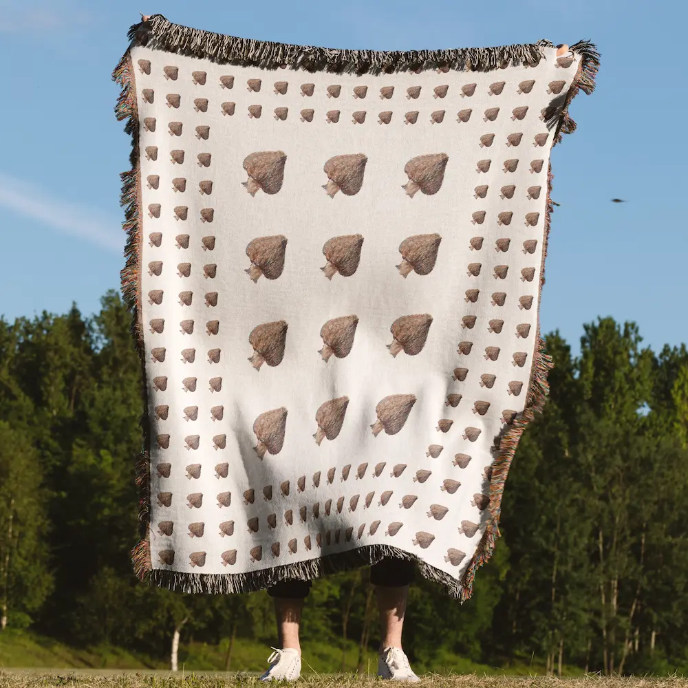 A person holding up the blanket outdoors with blue sky and green trees behind them, clearly showing the mushroom pattern and fringed edge.