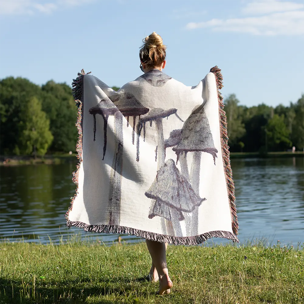 Person holding a shaggy mane mushroom blanket with oversized gray-purple mushroom illustrations and fringed edges beside a lake on a sunny day.